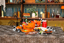 Halloween party table set with a black spiderweb table cover, orange Trick or Treat candy bowl, orange cutlery and cups, printed spider napkins and plates, mini cauldrons of candy corn and a skeleton garland