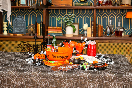 Halloween party table set with a black spiderweb table cover, orange Trick or Treat candy bowl, orange cutlery and cups, printed spider napkins and plates, mini cauldrons of candy corn and a skeleton garland